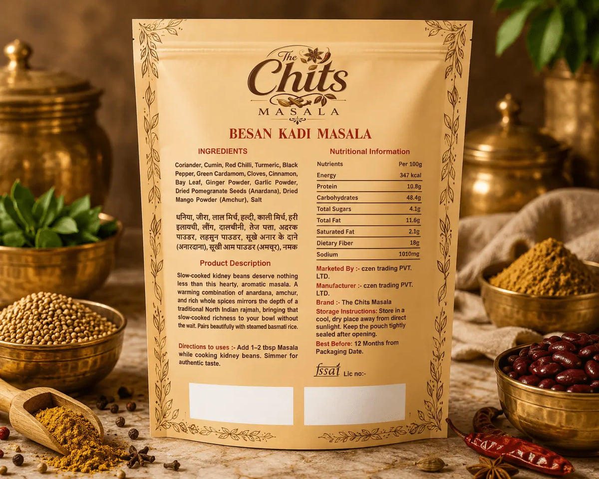 A standing printed card showing the back of a food product label for Besan Kadi Masala, photographed in a styled kitchen-like setting with spices, brass vessels, and a bowl of red chilies. The card is the main subject and contains product information, ingredients, nutrition facts, and directions.