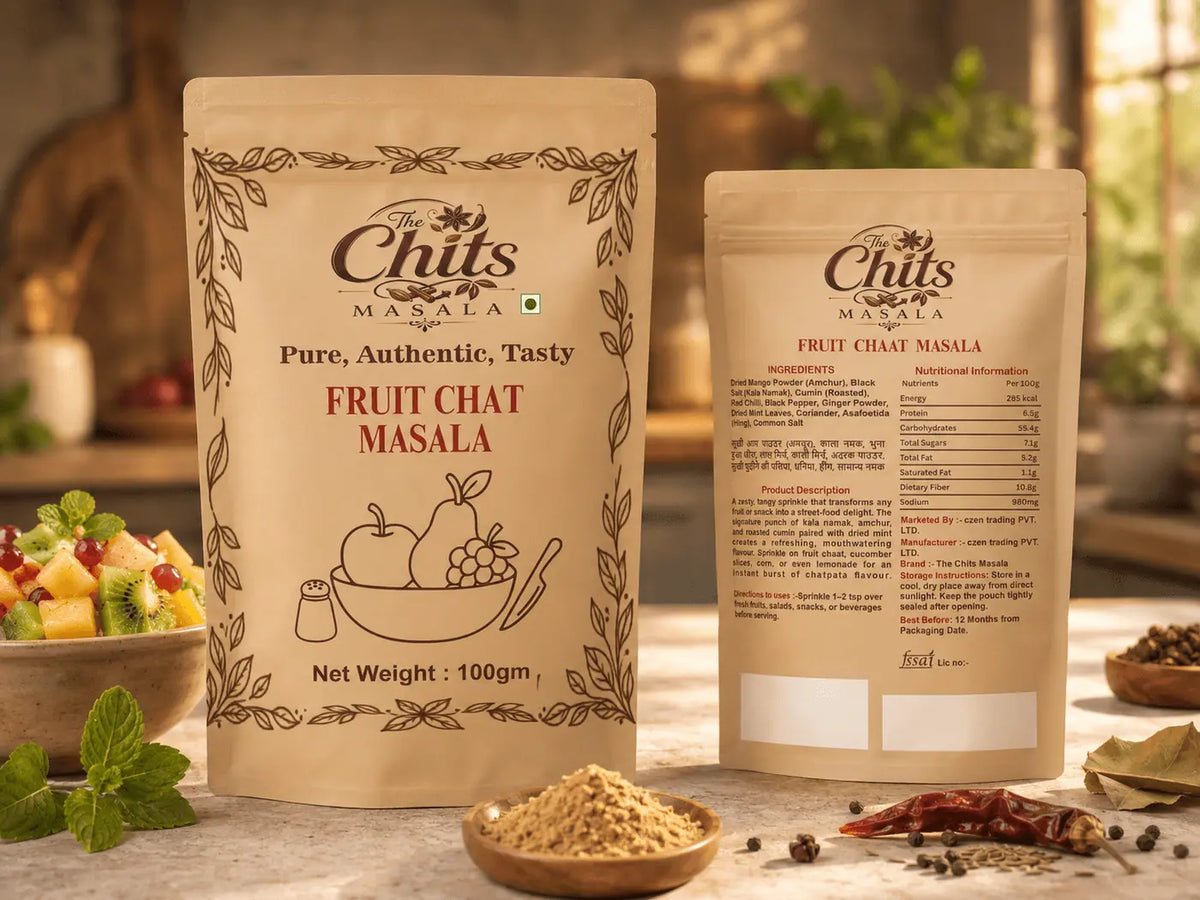 Two stand-up pouches of The Chits Masala Fruit Chat Masala displayed on a rustic wooden table with ingredients like dried red chilies, peppercorns, and mint leaves in the foreground.