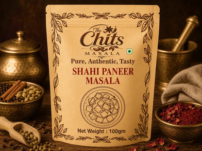 A 100g stand-up pouch of 'The Chits Masala Shahi Paneer Masala' centered on a rustic wooden table surrounded by spices, bowls, and a mortar and pestle.